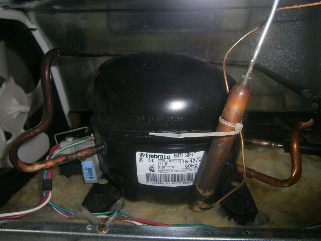 Close-up of a household refrigerator compressor with copper refrigerant tubing at the back of a fridge